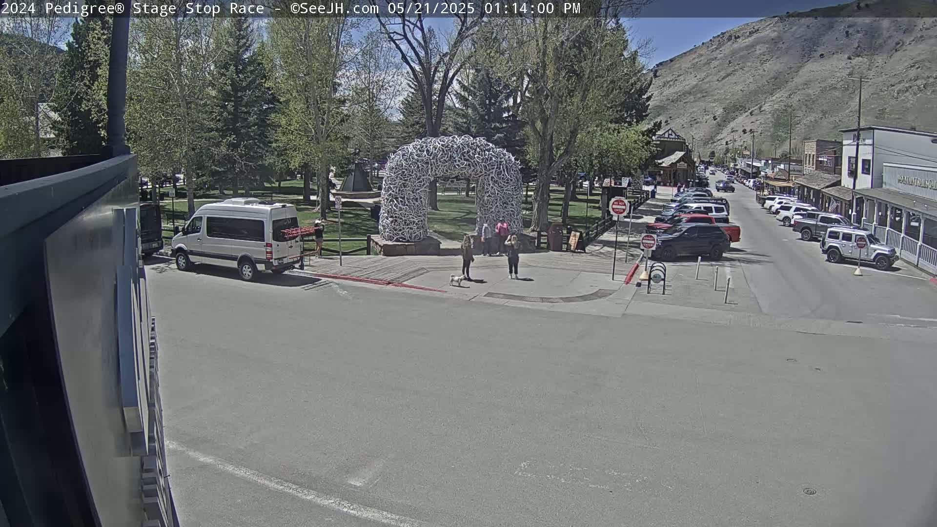 A sunny day shows a town square with a large arch made of antlers, several parked cars lining the street, and a few pedestrians near the arch.