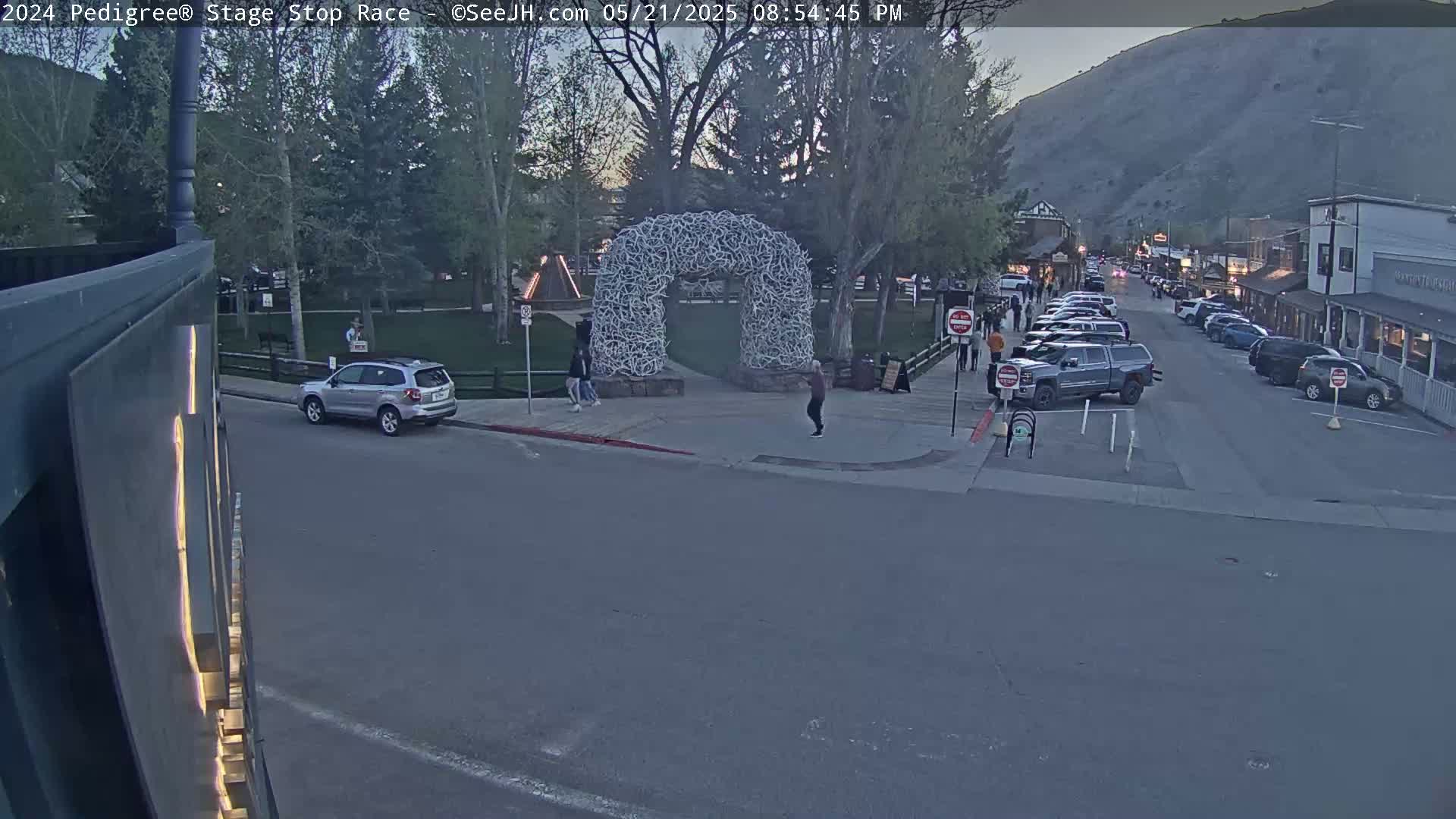 A street scene at dusk shows several cars parked along the side of a road, with a large archway made of antlers in a park-like area across the street.