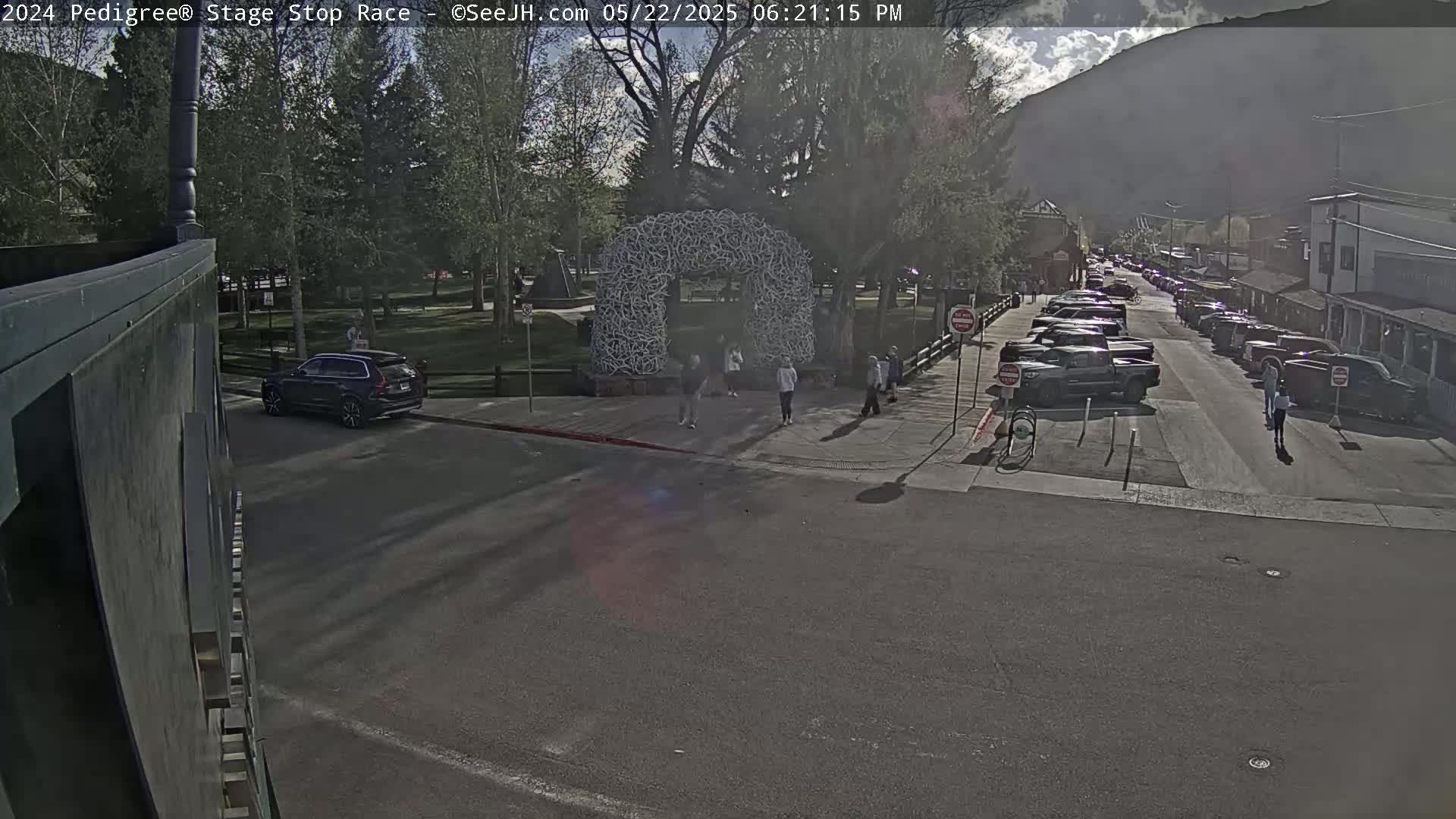 A partly cloudy afternoon shows a street scene with parked cars lining both sides, pedestrians crossing near a large, white, arch-like structure in a park-like area, and a mountain visible in the background.