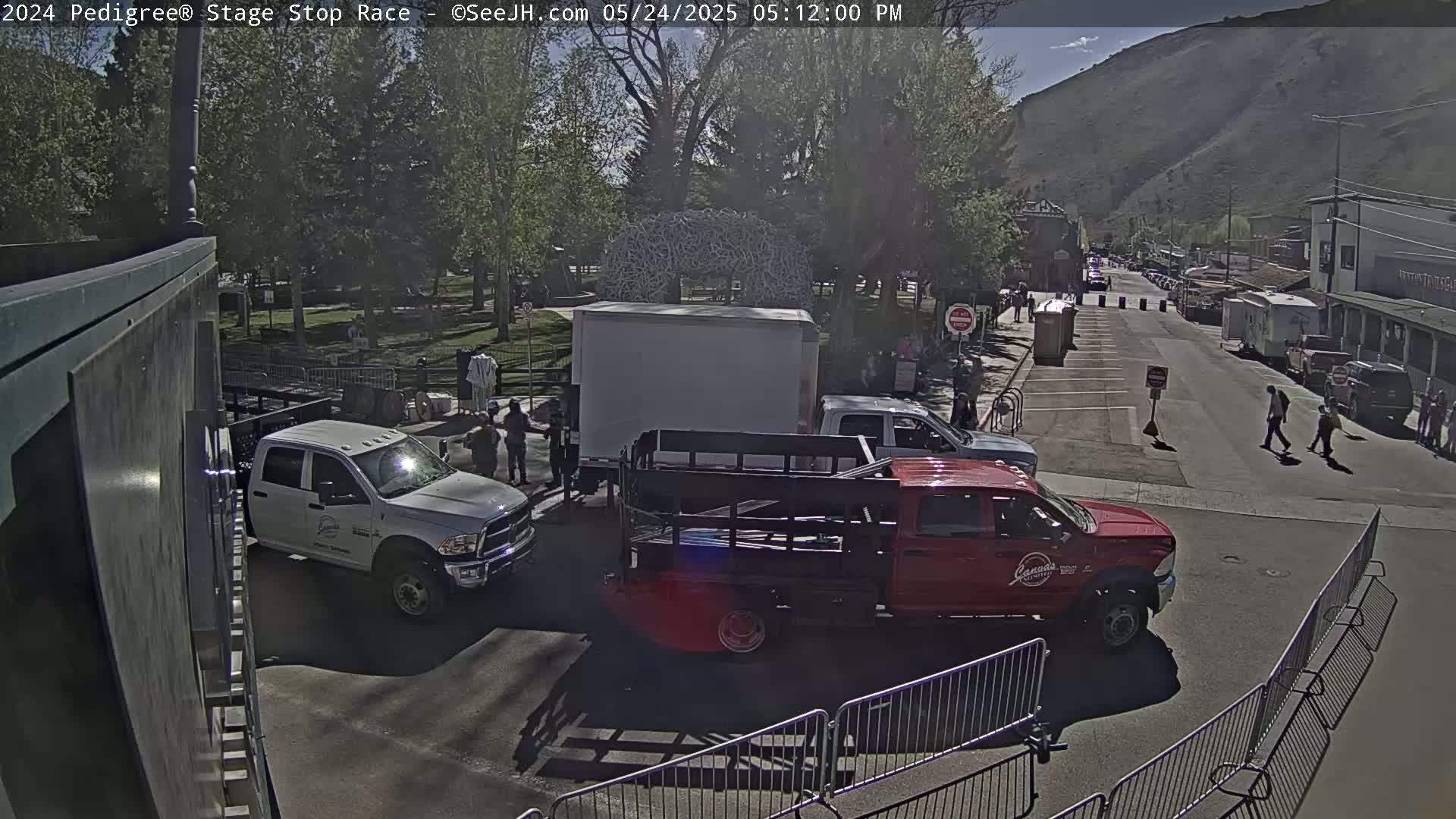A sunny day reveals a street scene with several trucks parked near a large white box truck, people milling about, and a mountain in the background.