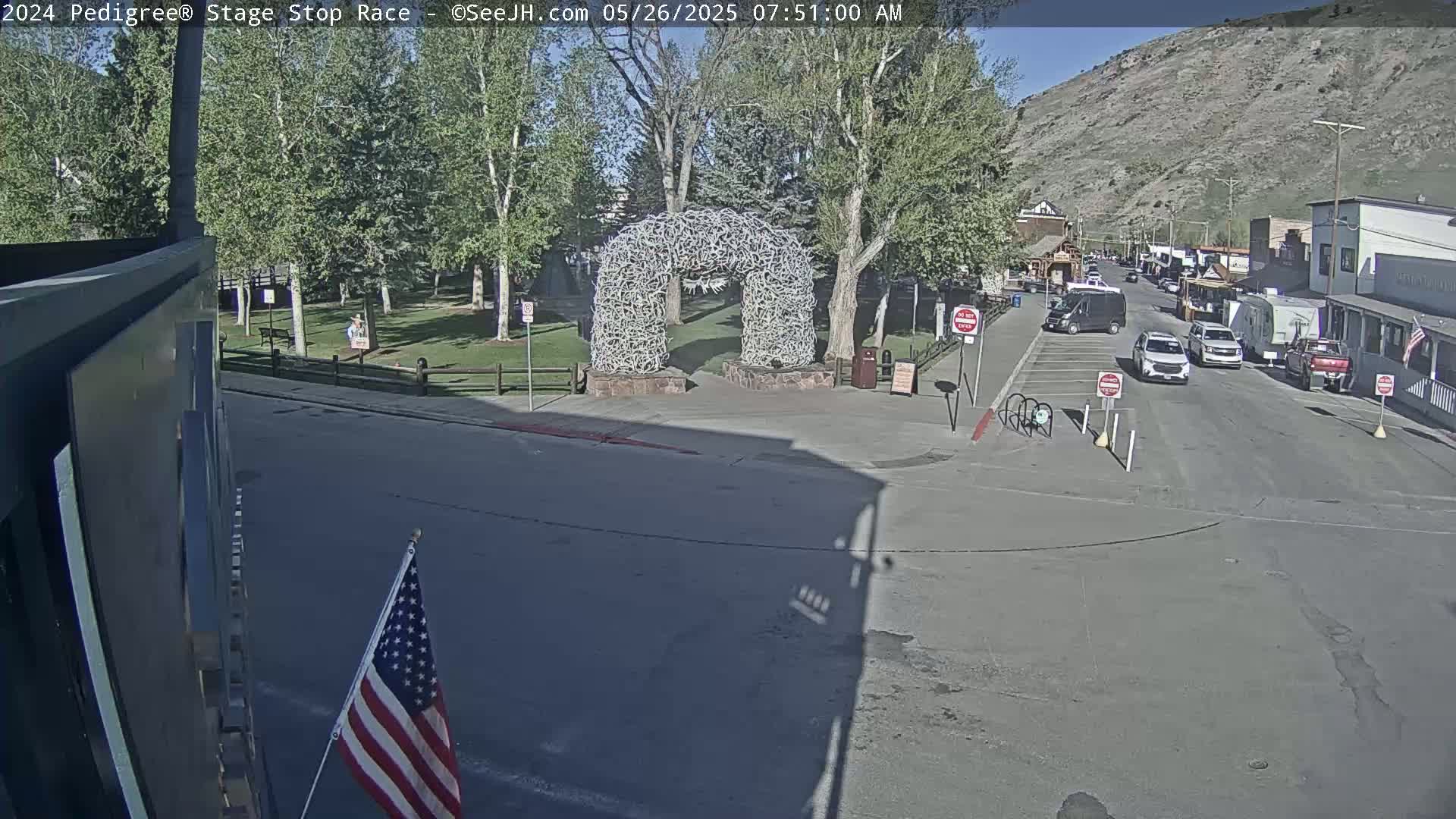 A street scene on a sunny day shows a town with several vehicles parked along the street, a large antler archway in a nearby park, and a small American flag displayed.