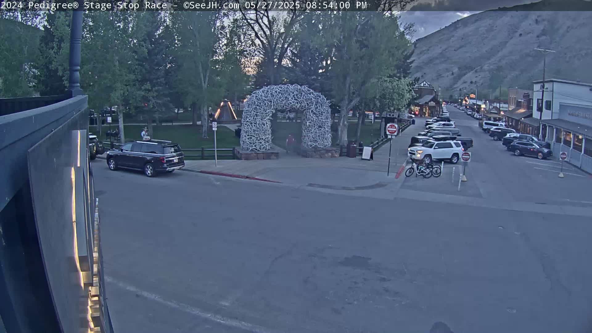 A street scene at dusk shows cars parked along a street in front of buildings, with a large antler arch in a park across the street.