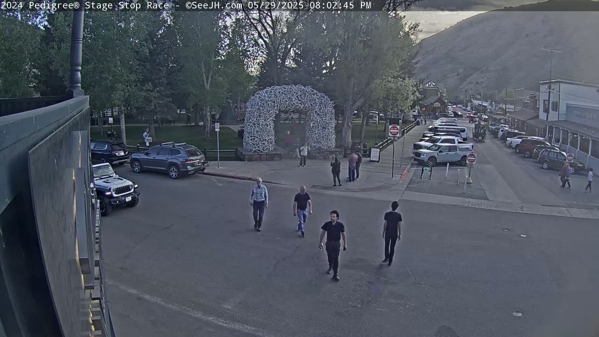 Several people walk near parked cars and a large antler archway in a town under a partly cloudy sky.