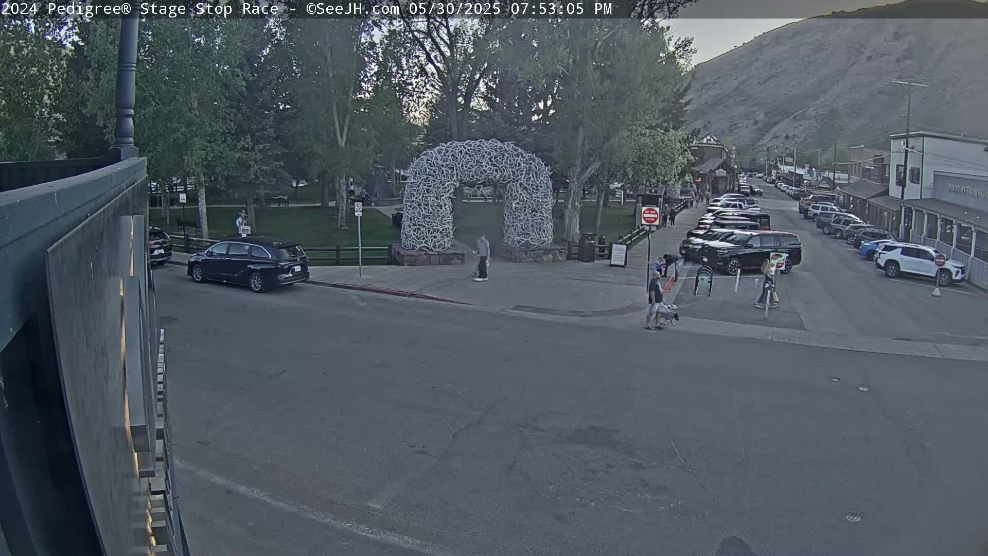 A street scene at dusk shows a town square with a large antler archway, several parked cars, and a few pedestrians; a mountain is visible in the background.