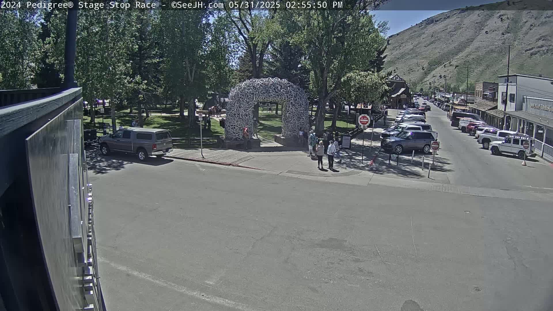 A sunny day in a town with a park featuring an arch made of animal horns, a street lined with parked cars, and pedestrians near the park entrance.