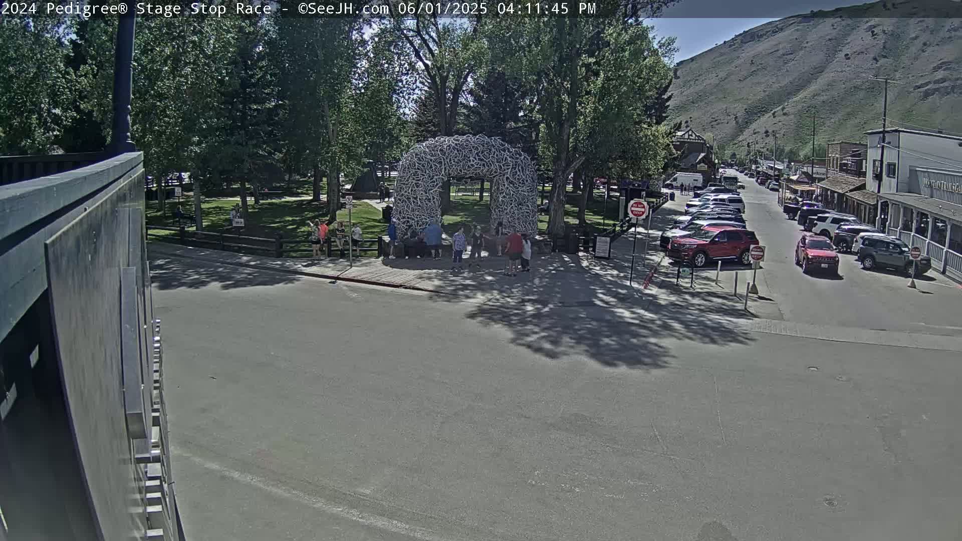 A large antler archway stands in a town square with people gathered around it, under a sunny sky, with parked cars lining a street leading to a mountain in the background.