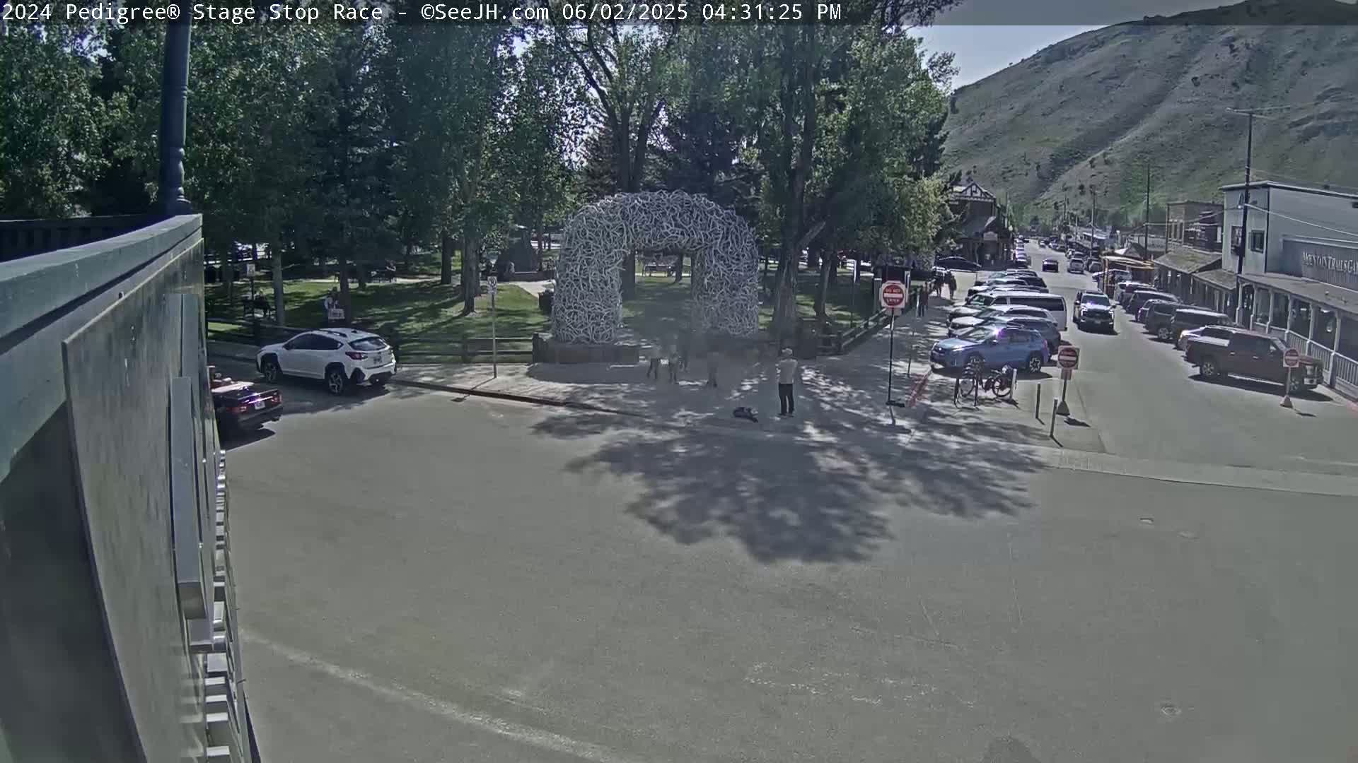 A sunny day reveals a street scene with parked cars, pedestrians near a large antler archway in a park-like setting, and a mountain visible in the background.