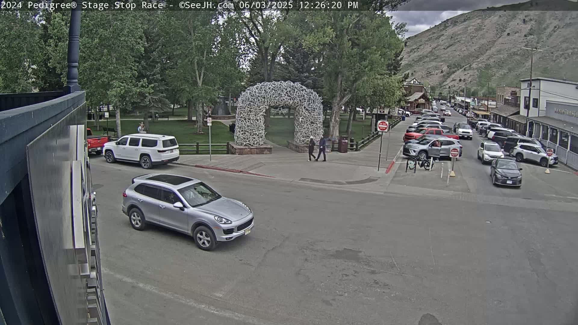 A silver SUV drives past a large antler archway in a park, with several cars parked along a street in a town nestled against a mountain, under partly cloudy skies.