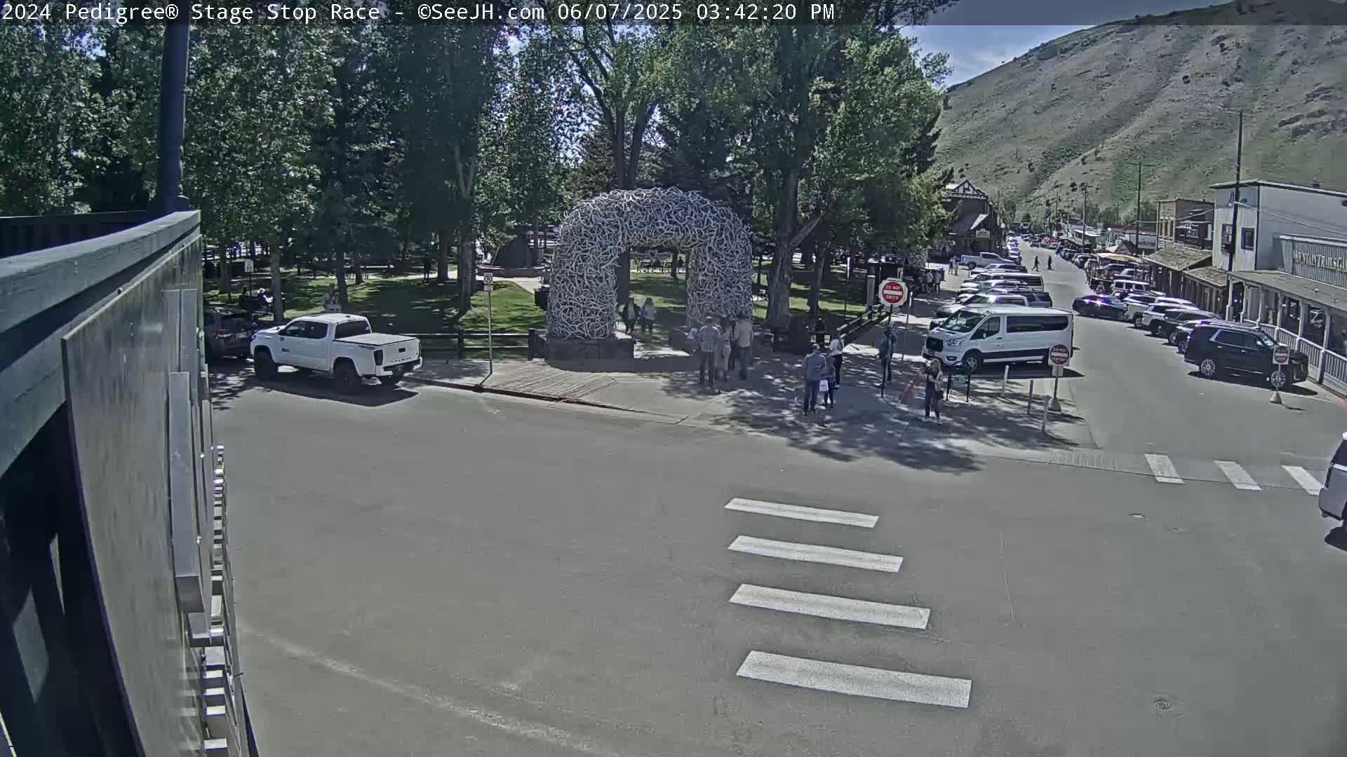 A sunny day scene shows a town square with parked cars, pedestrians near a large antler arch, and a white pickup truck at an intersection.