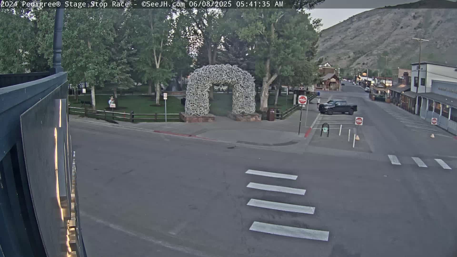 A dark-colored pickup truck is parked near a crosswalk in a town square under a twilight sky, with an antler archway in the background.