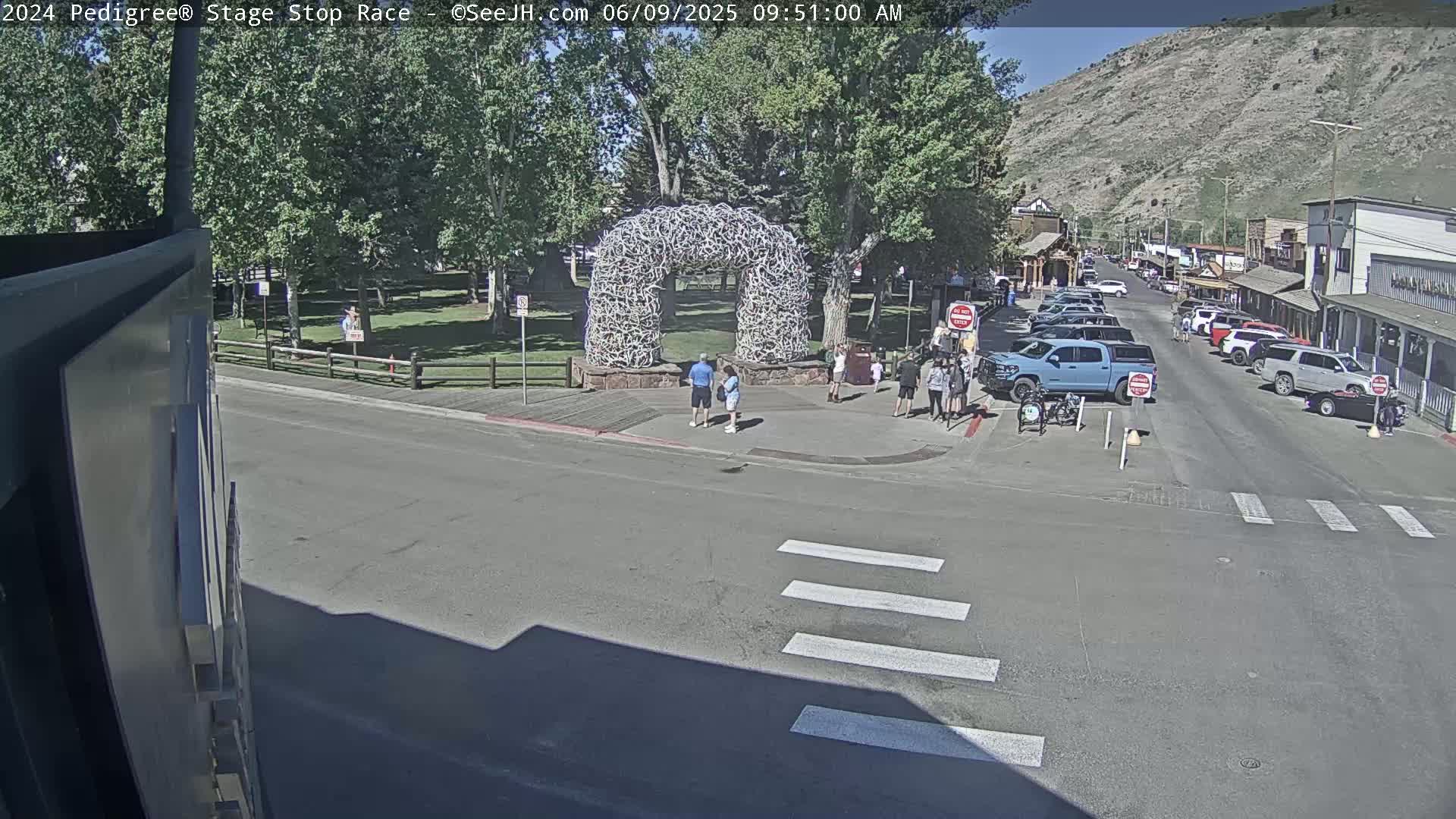 A sunny street scene shows a crosswalk, several parked cars, pedestrians near a large antler arch, and a background of buildings and a mountain under a clear sky.