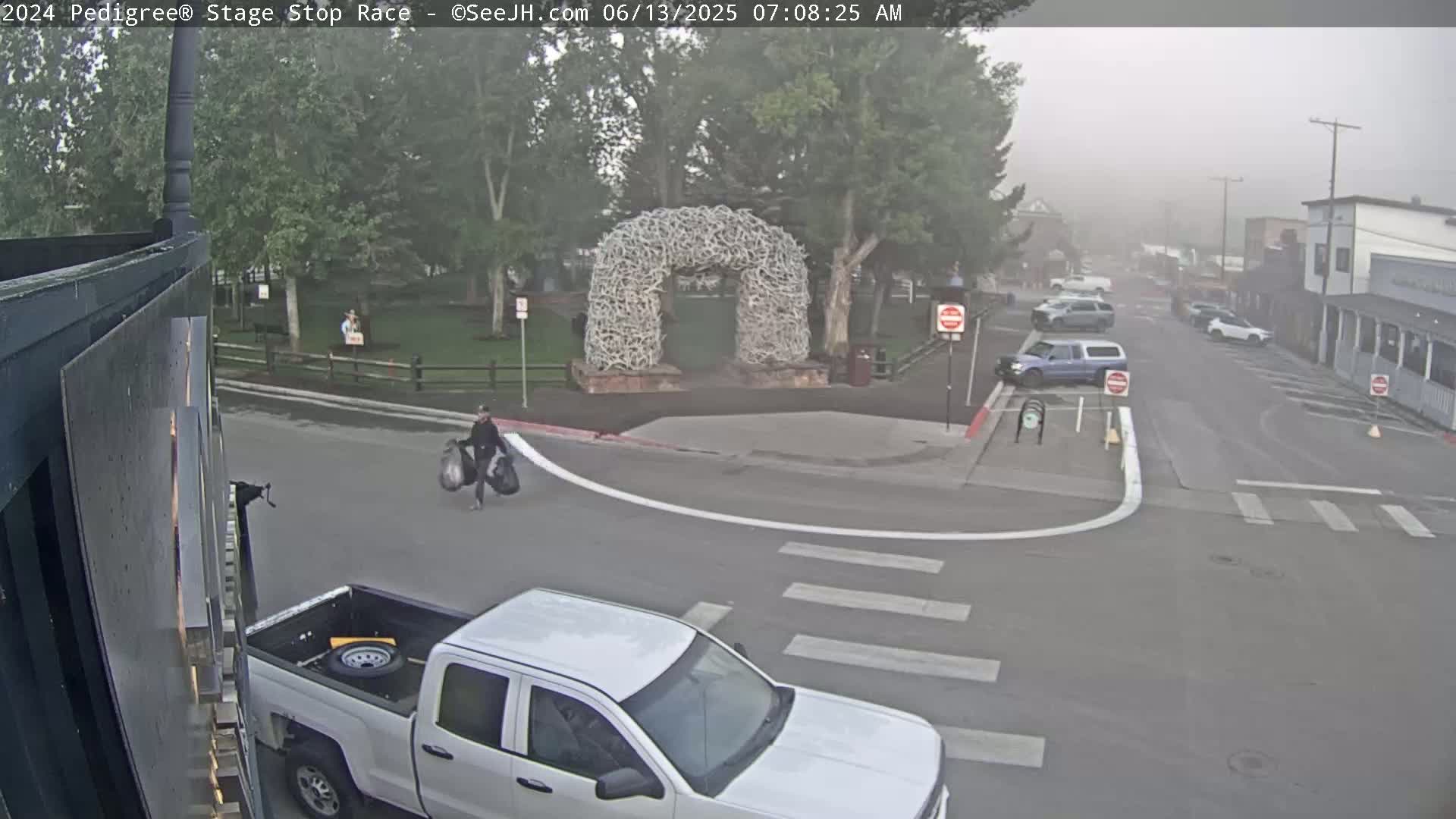 A person carries bags near a white pickup truck at a street intersection in a foggy town, with a large antler arch visible in the background.