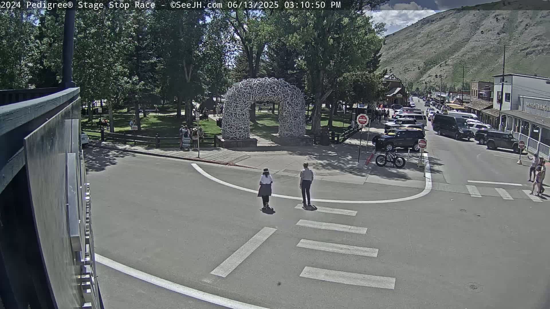 Two people cross a street in front of a large arch made of antlers, near parked cars and buildings on a sunny day.