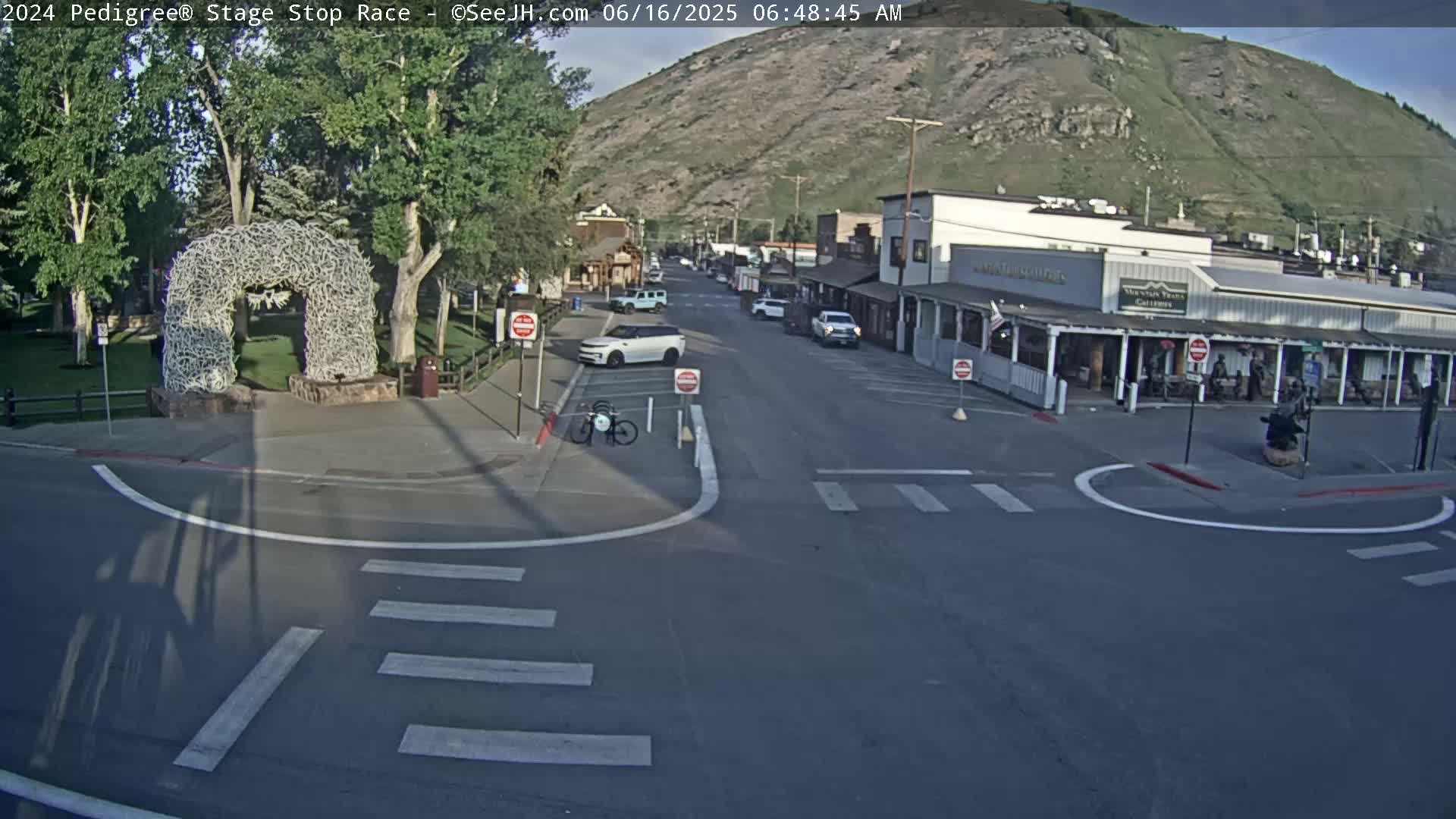 A mostly empty street in a town nestled against a mountainside on a sunny morning, featuring a large antler archway in a nearby park.