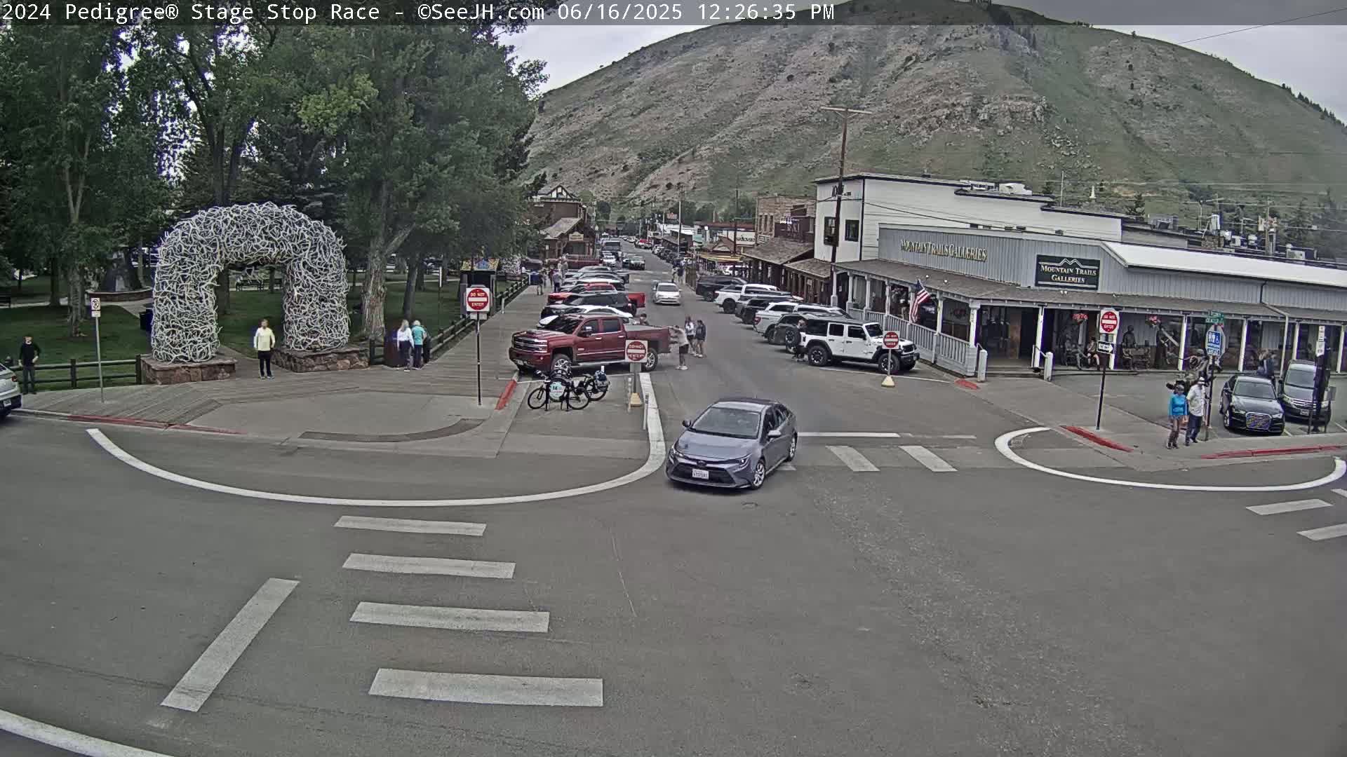 A street scene in a town nestled against a mountain, with parked cars and pedestrians near shops under a clear sky.