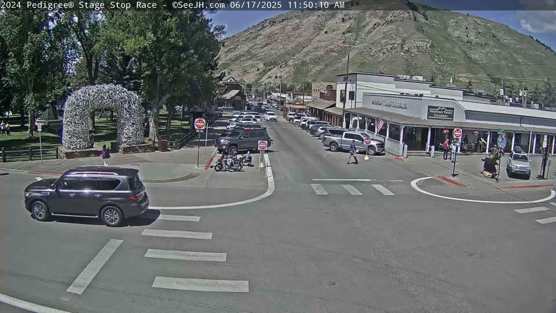 A sunny day shows a street intersection in a town nestled against a mountain, with several cars parked along the sides of the street and a few pedestrians walking.