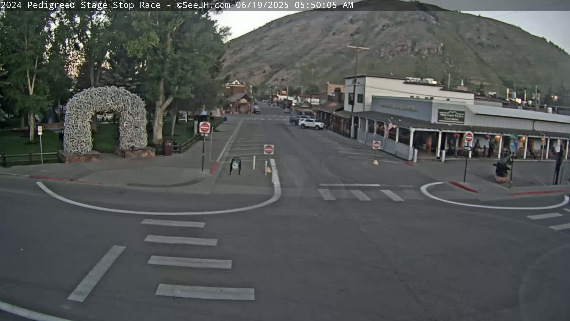 A street intersection in a town at the base of a mountain under a clear, early morning sky features a large antler arch in a nearby park.