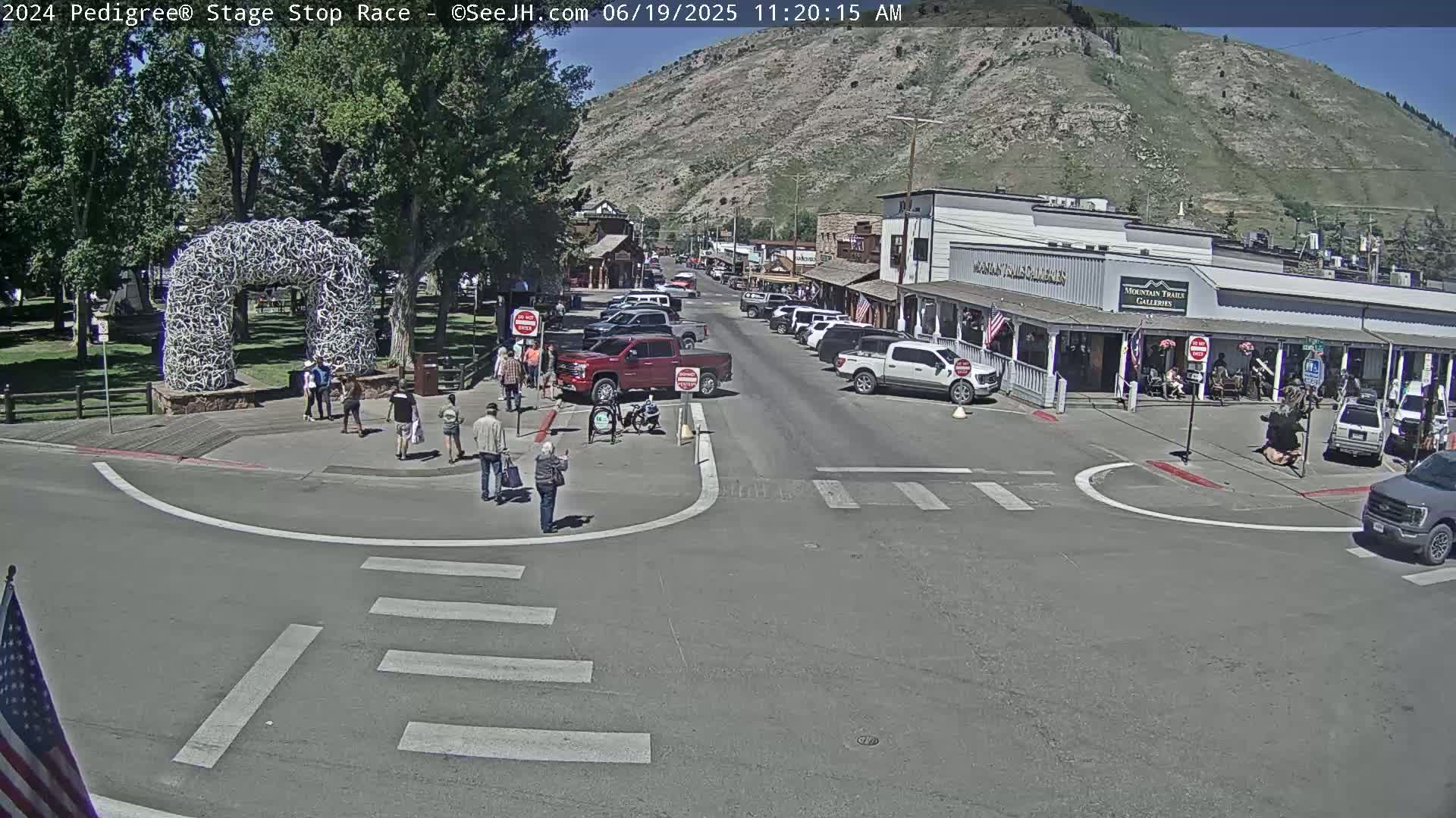 A sunny town square with pedestrians and parked cars, under a clear sky and near a mountain.