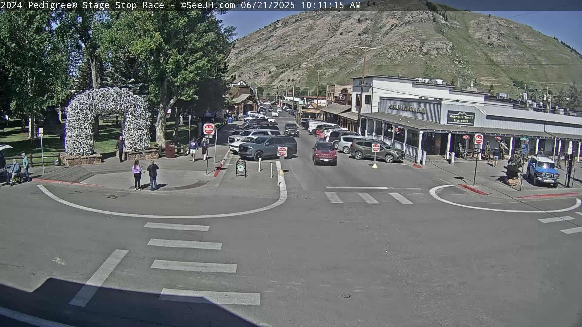 A sunny day reveals a town square with a large antler arch, several parked cars, a few pedestrians, and buildings lining a street against a mountain backdrop.