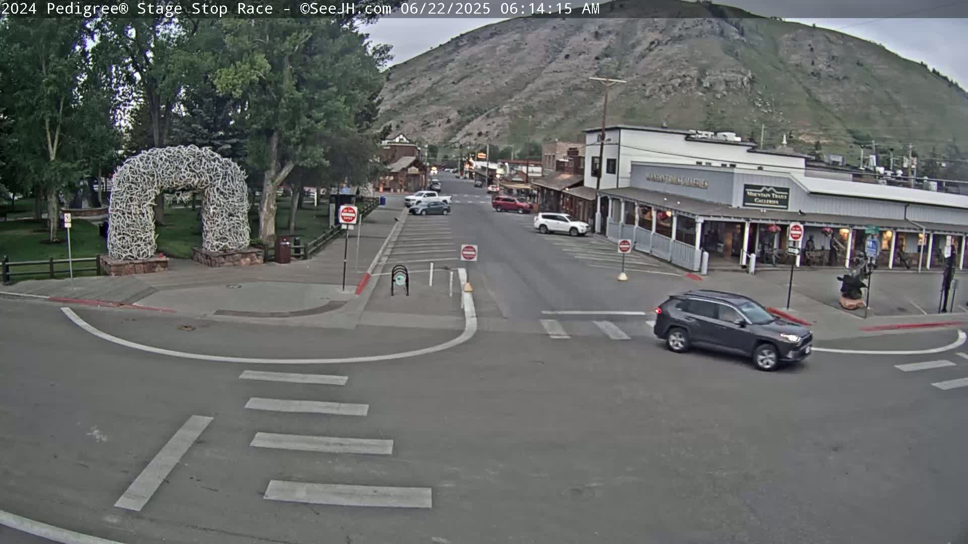 A grey SUV drives past a large antler archway in a town nestled against a mountainside on a partly cloudy morning.