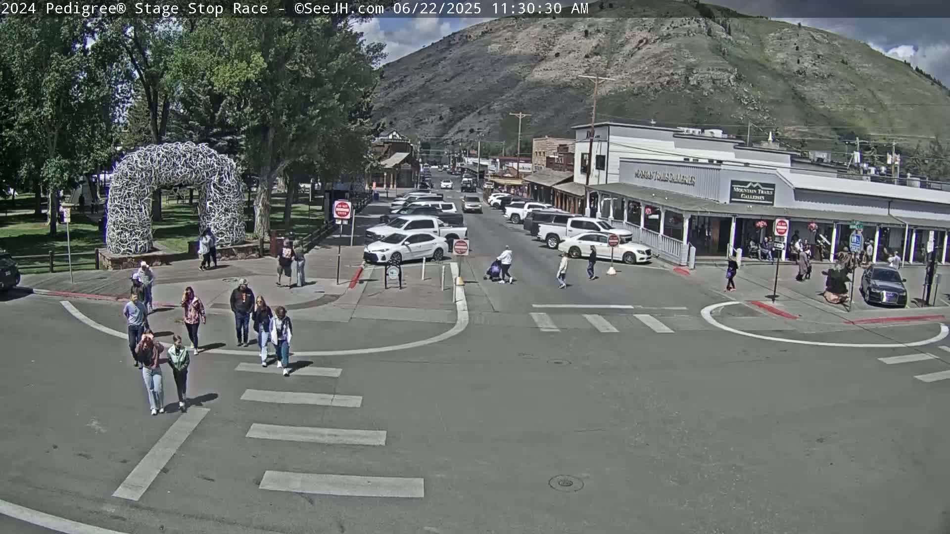 Pedestrians cross a street intersection in front of shops and buildings nestled against a mountainside on a sunny day.