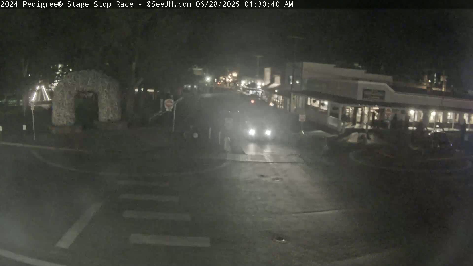A nighttime street scene shows a vehicle driving past a building with people outside, under foggy or misty conditions.