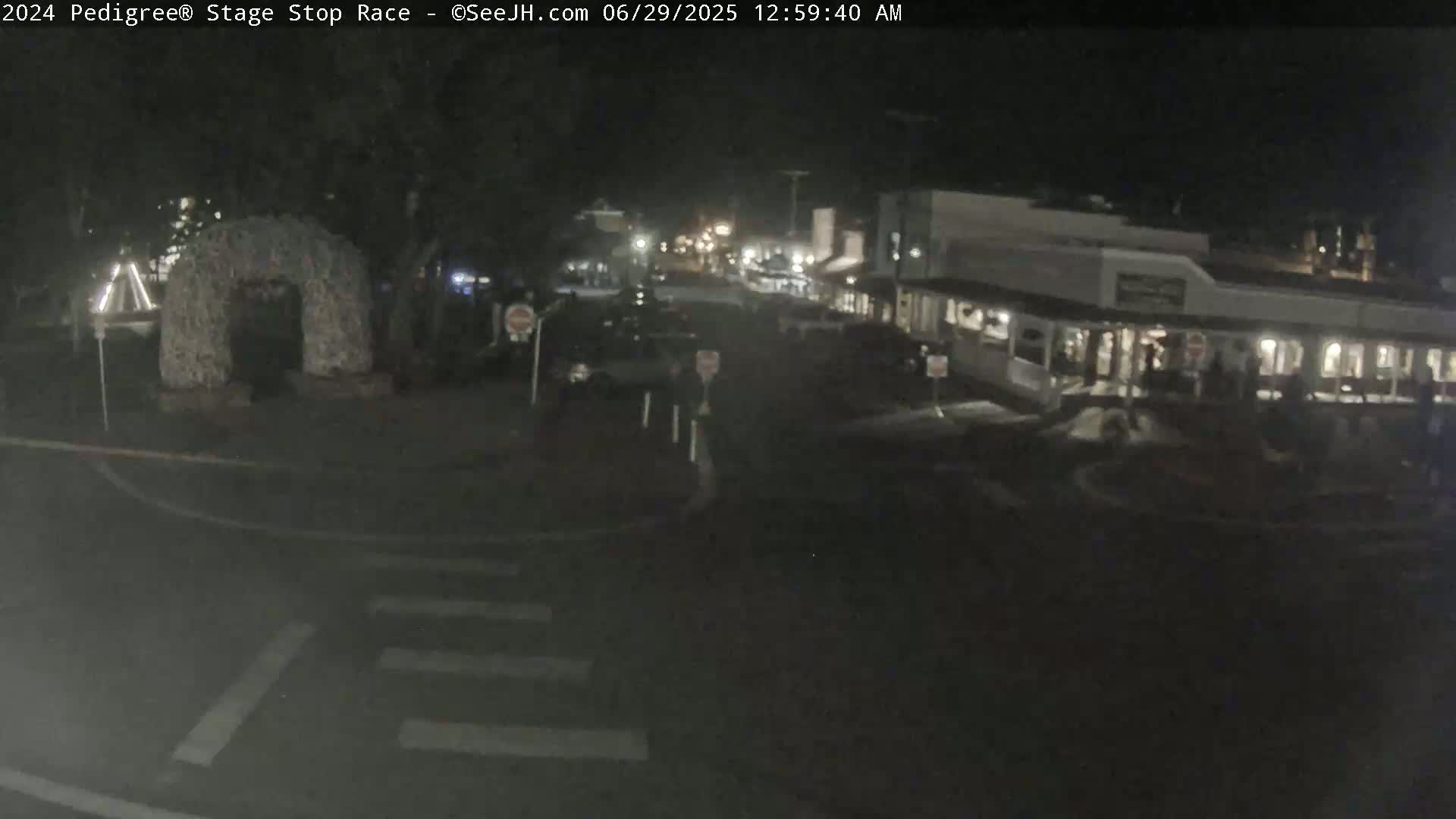 A nighttime, dimly lit street scene shows parked cars, a few pedestrians, and a stone archway.