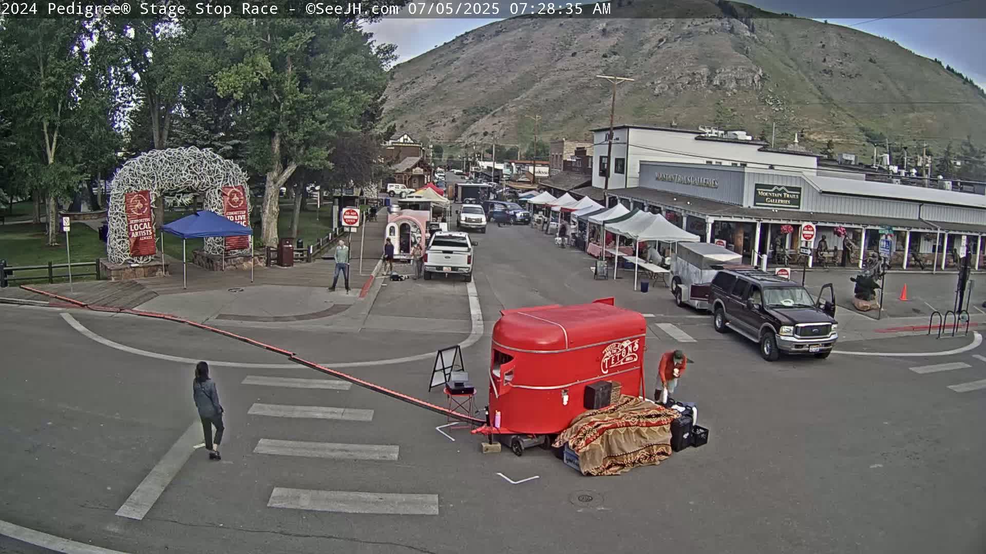 A street scene shows a small town with several market stalls and vendors, a red food truck, and pedestrians, all under a mostly clear sky in front of a mountain.