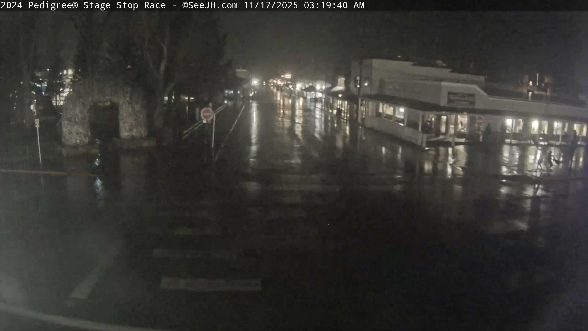 A wet street in a town at night reflects building and streetlights on its slick pavement, indicating rainy conditions.
