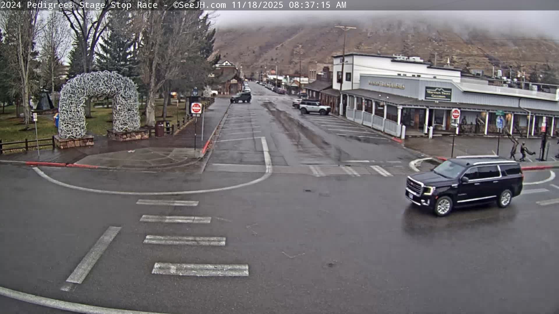 A wet street in a town at night reflects building and streetlights on its slick pavement, indicating rainy conditions.