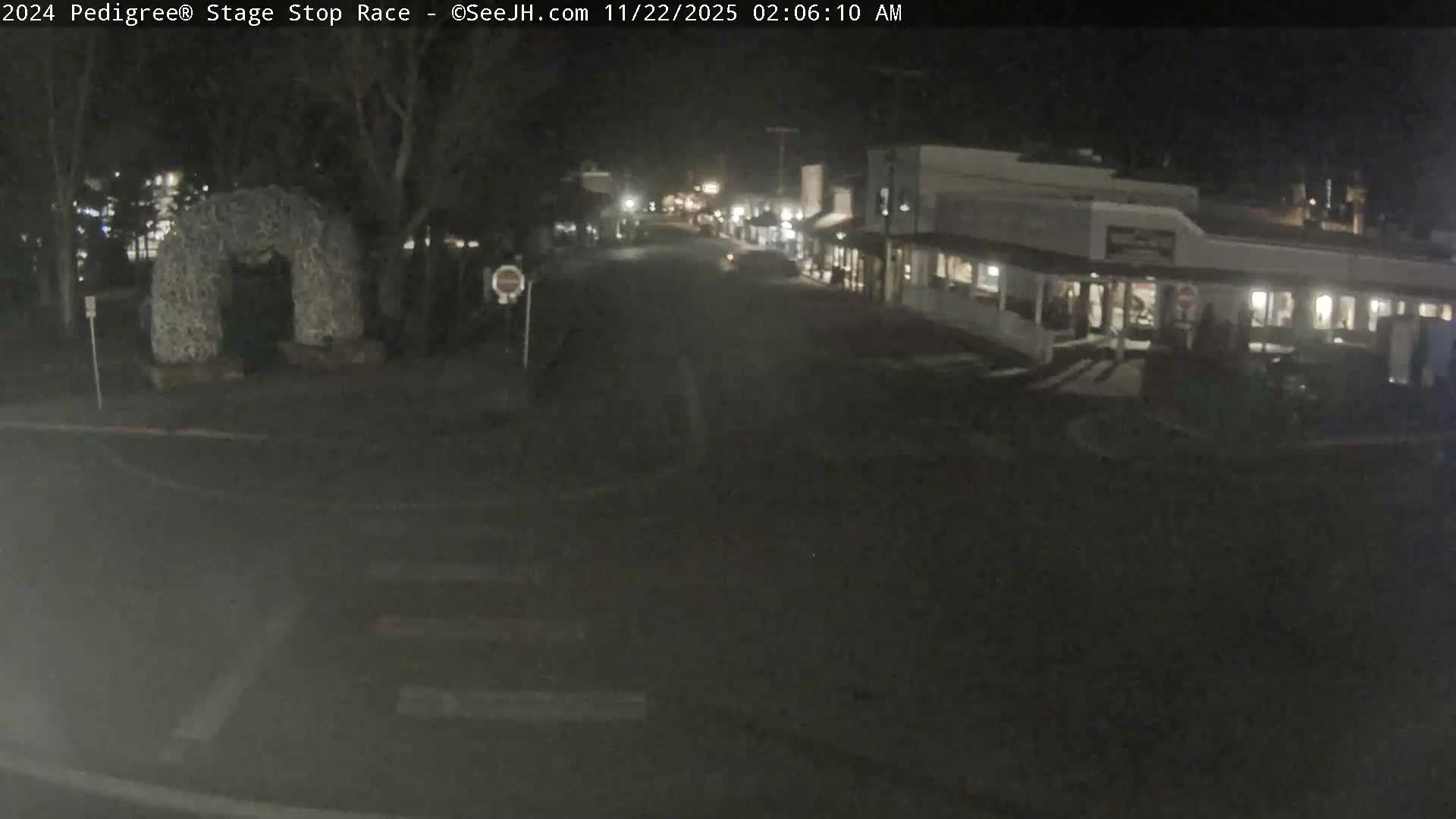 This nighttime image captures a quiet street scene featuring a large, textured archway on the left, faint crosswalk markings on the dark asphalt, and a row of illuminated buildings stretching into the distance on the right under a clear sky.