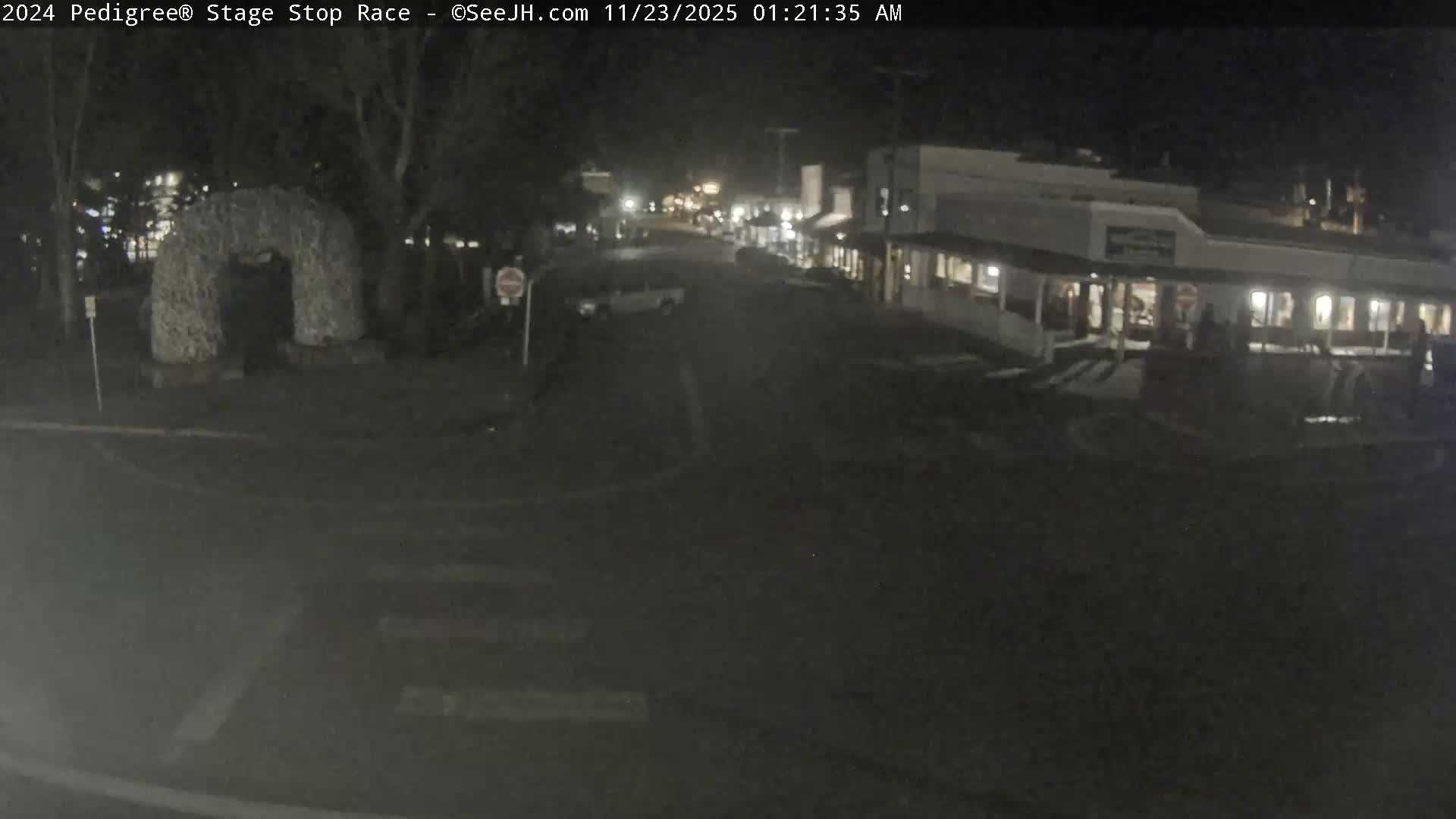 The image shows a dark, clear night scene of a quiet street in a town, featuring a decorative archway on the left, an empty street with crosswalks, and illuminated buildings lining the right side.