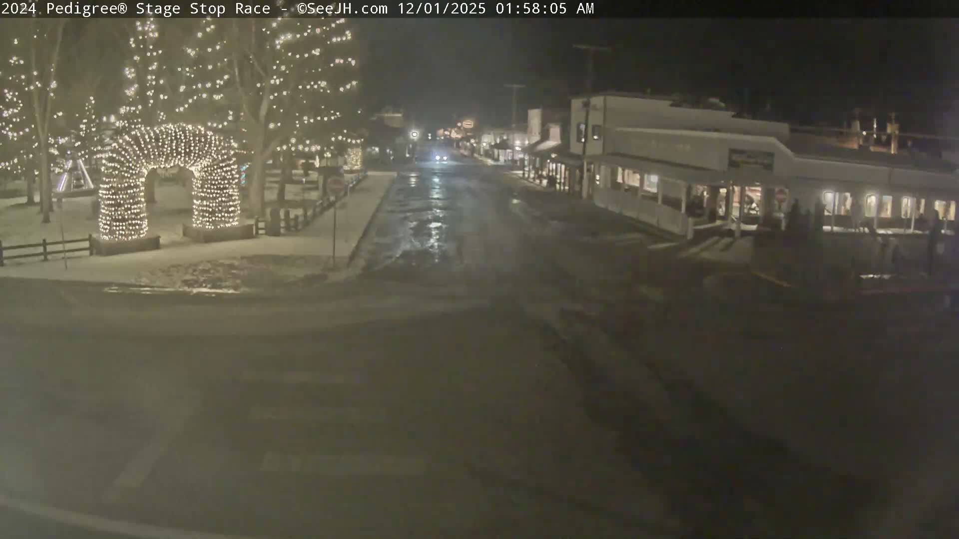 A wet and snowy town street is brightly illuminated at night by festive lights on trees and a prominent archway on the left, with a row of storefronts lighting up buildings on the right.