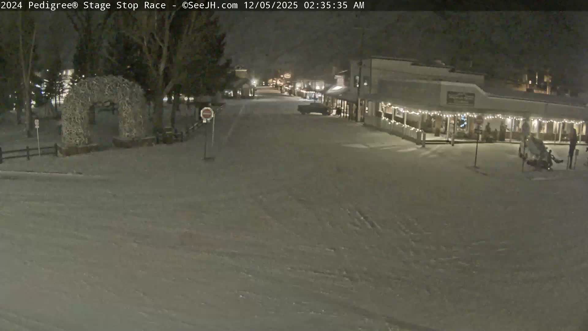 A quiet, snow-covered street at night, lined with buildings illuminated by string lights on the right and featuring a prominent antler archway on the left, is experiencing a gentle snowfall.