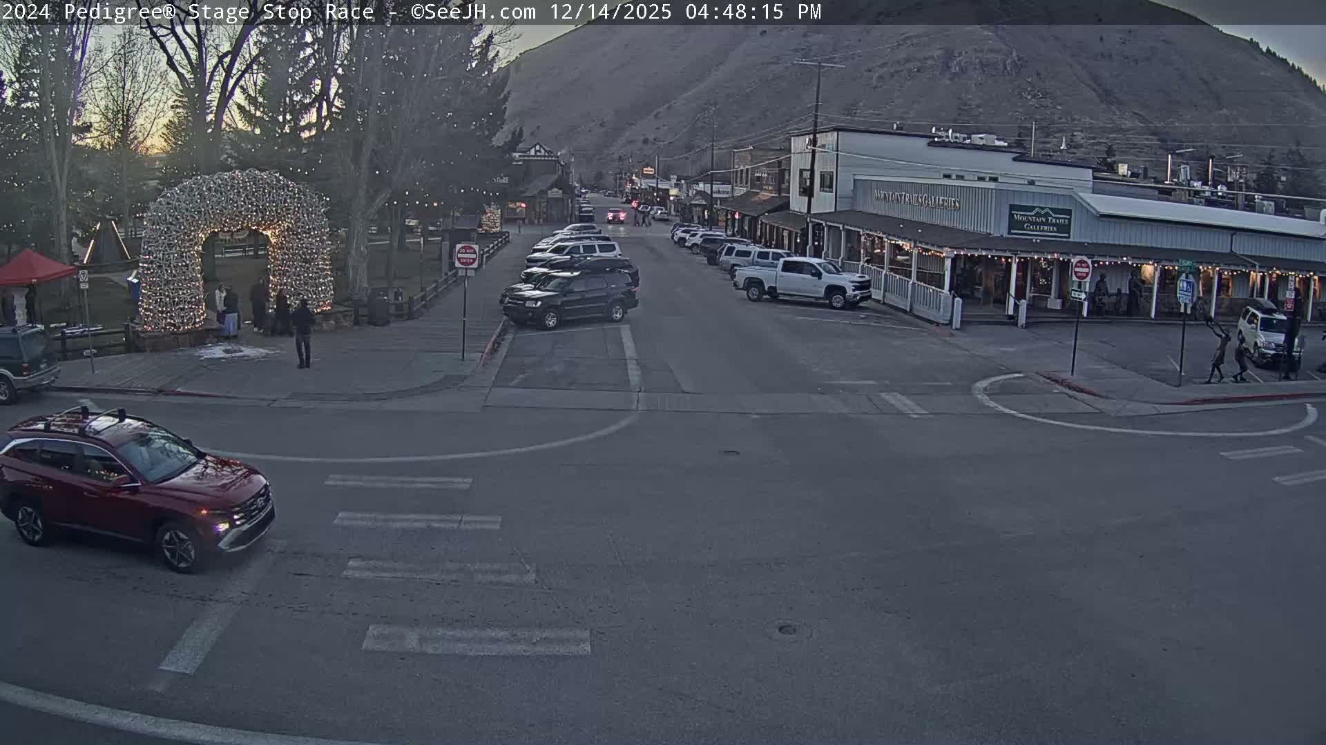 A quiet, snow-covered street at night, lined with buildings illuminated by string lights on the right and featuring a prominent antler archway on the left, is experiencing a gentle snowfall.