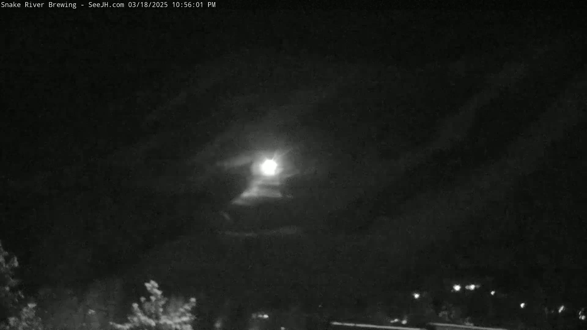 A bright moon shines through a partly cloudy night sky above a dark landscape with a few distant lights.