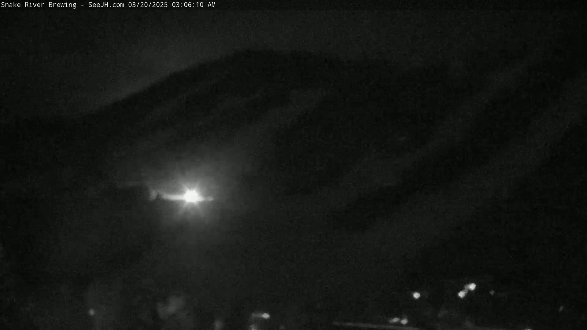 A nighttime view of a dark mountain range under a cloudy sky, illuminated by a bright light source in the mid-ground.
