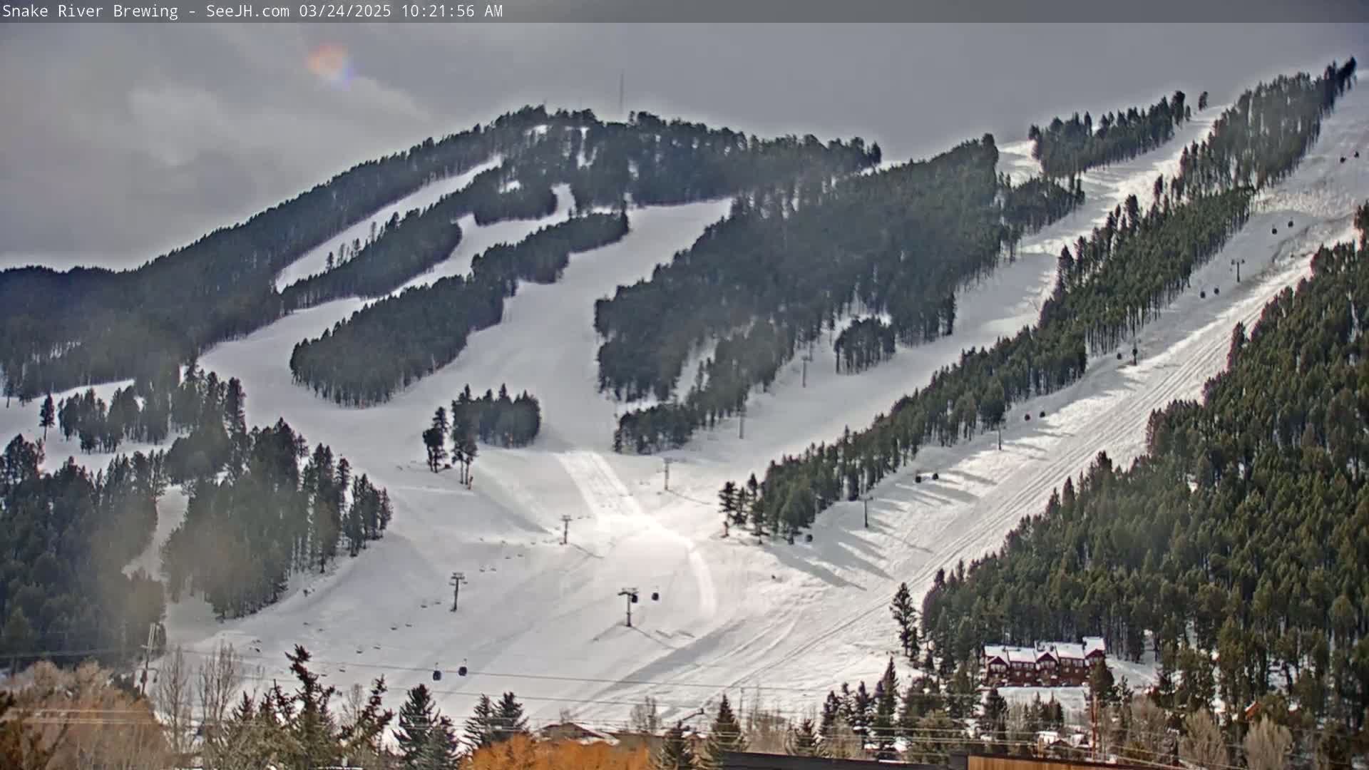 A snow-covered ski resort with several runs winding through evergreen trees under a cloudy sky.