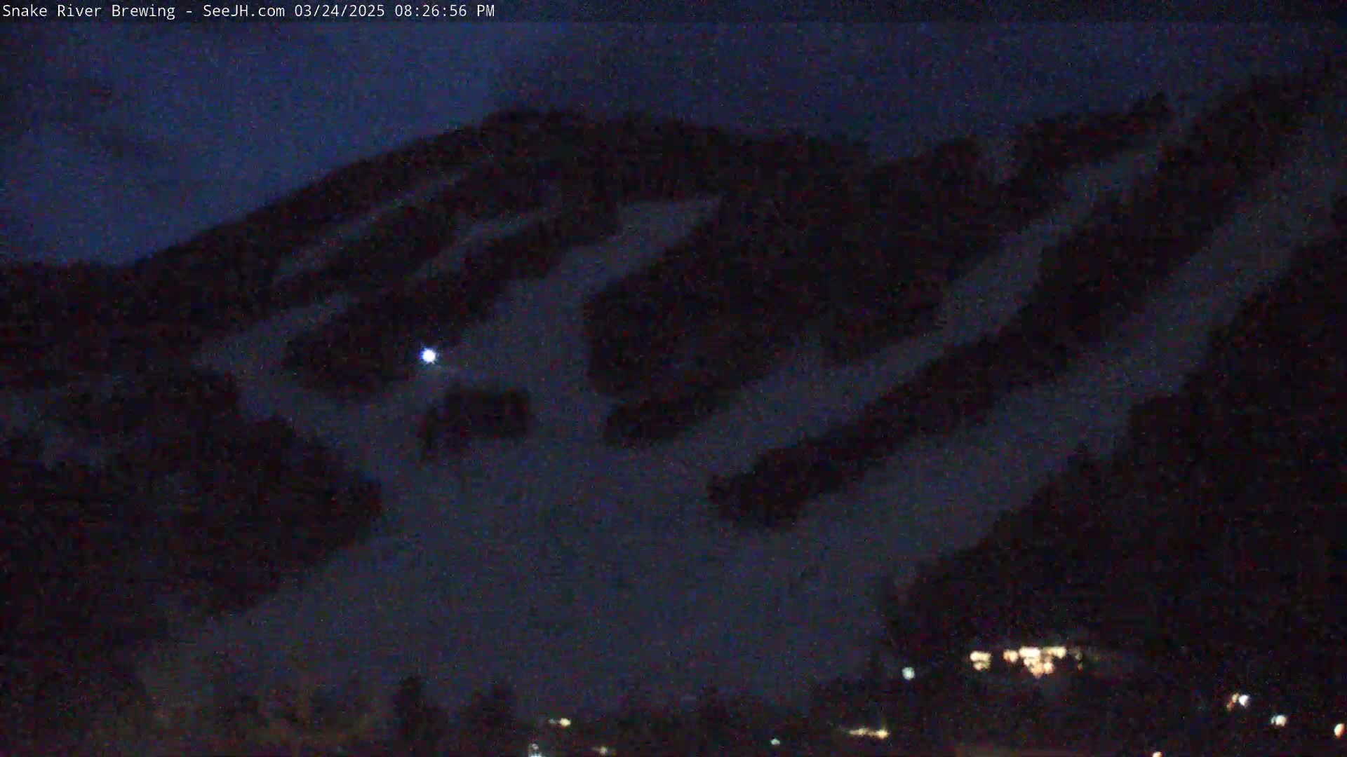 A dark, nighttime view of a snow-covered hillside under a mostly clear sky, with a few lights visible in the distance.