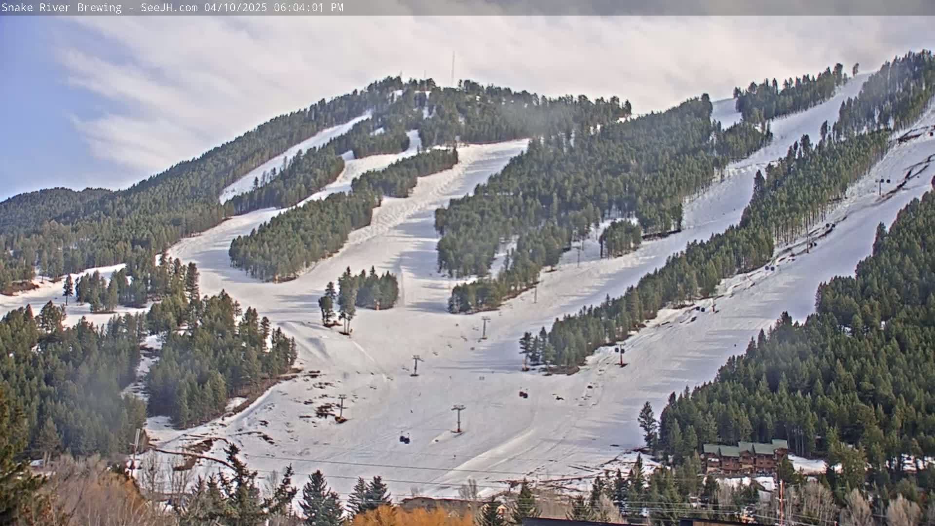 A snow-covered ski resort with numerous ski runs winding through evergreen trees under a partly cloudy sky.