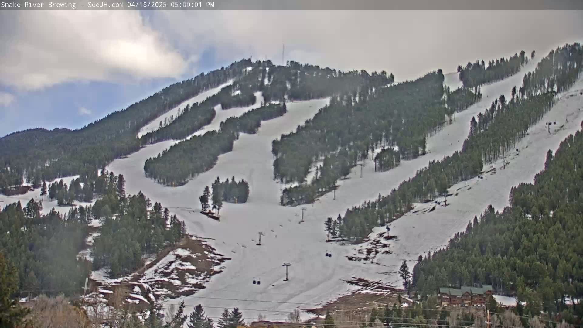 A snow-covered ski slope with several ski lifts and evergreen trees under a partly cloudy sky.