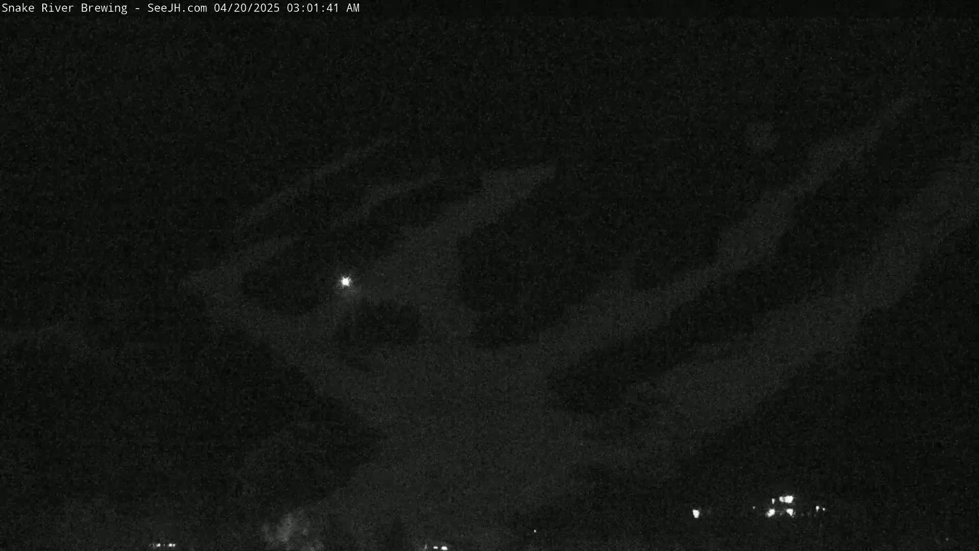 A nighttime image shows a mostly cloudy sky with a bright light source visible, and a few faint lights on the ground in the distance.
