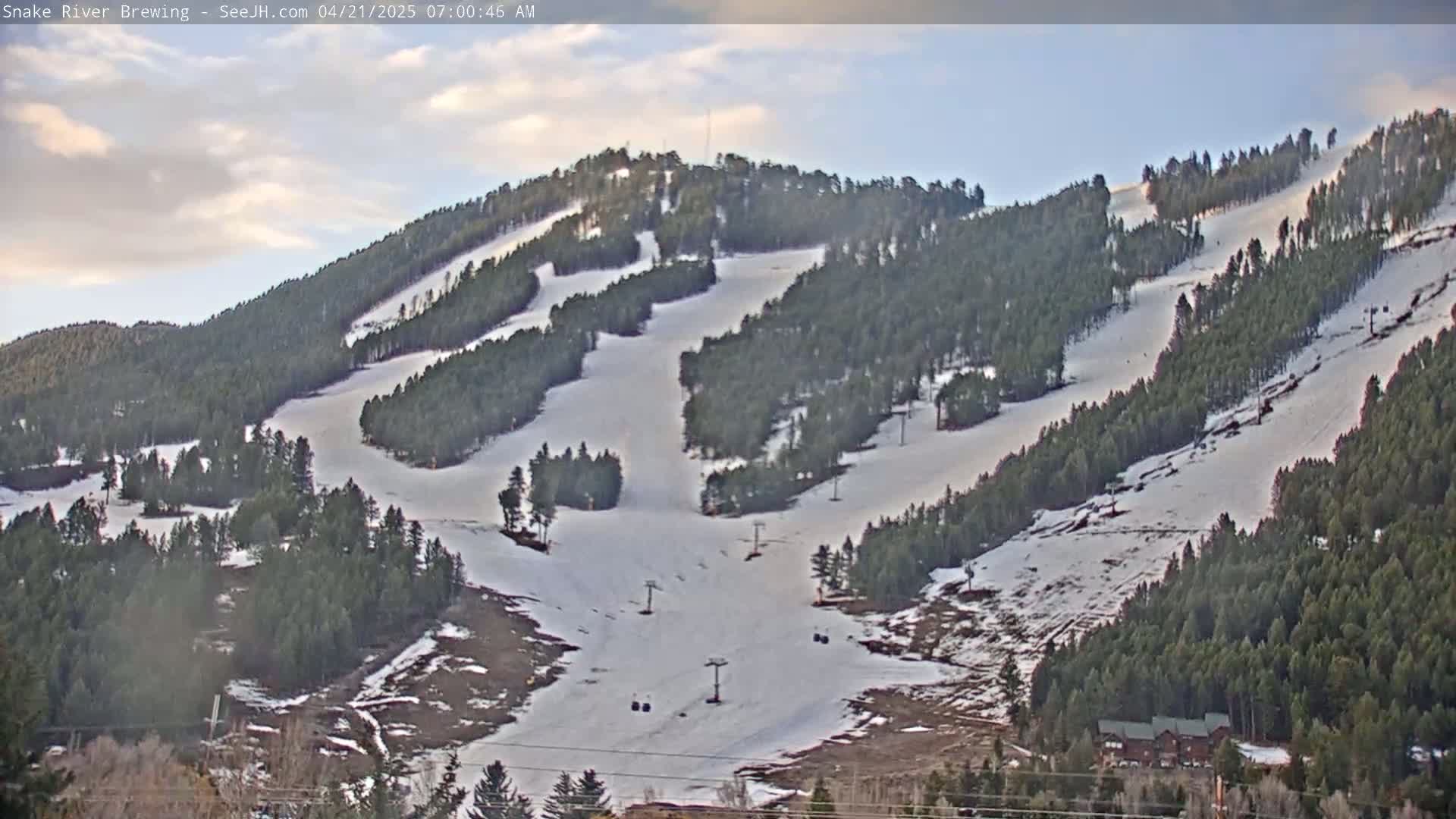 A snow-covered ski slope on a partly cloudy morning, with evergreen trees lining the mountainside and several ski lifts visible.