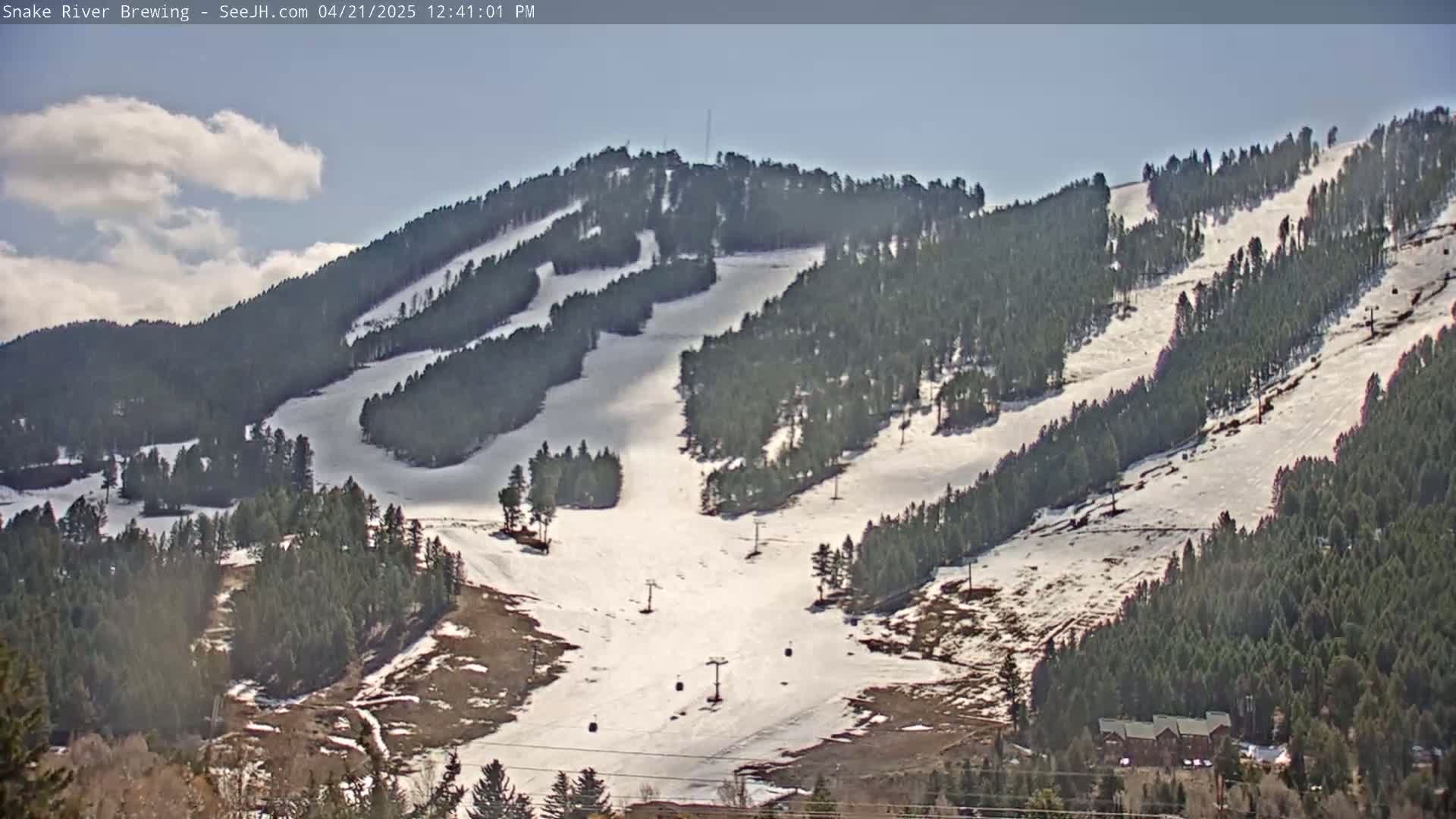 A snow-covered ski slope on a partly cloudy day, with evergreen trees lining the mountainside and ski lifts visible.