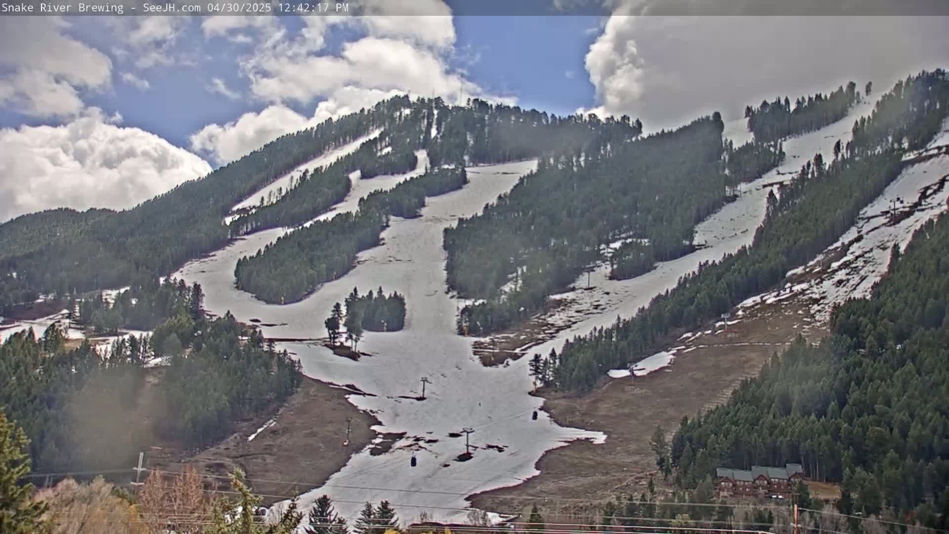 A partially snow-covered ski slope, dotted with evergreen trees, is visible under a partly cloudy sky.
