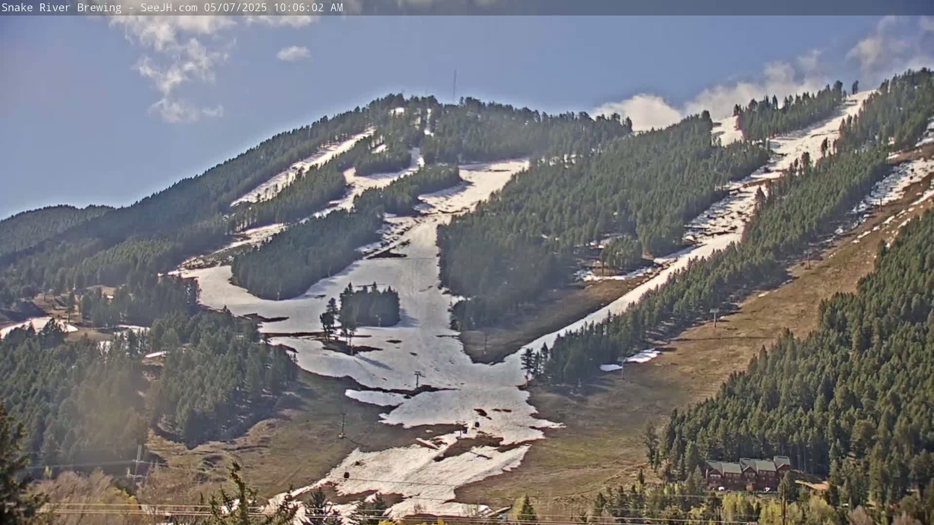 A mostly sunny day shows a mountainside ski slope with patches of remaining snow, surrounded by evergreen trees.