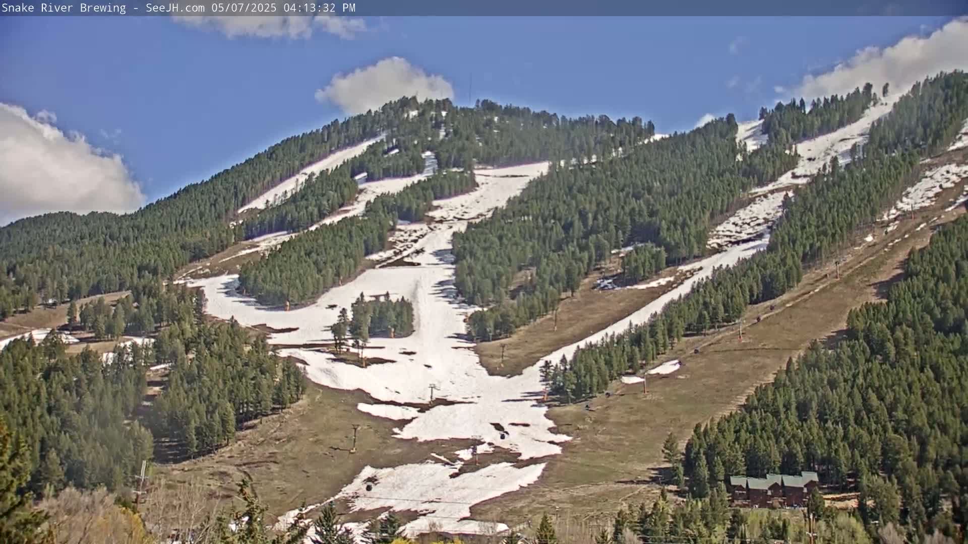 A partly sunny day reveals a snow-dusted mountainside with numerous ski runs winding through a dense evergreen forest.