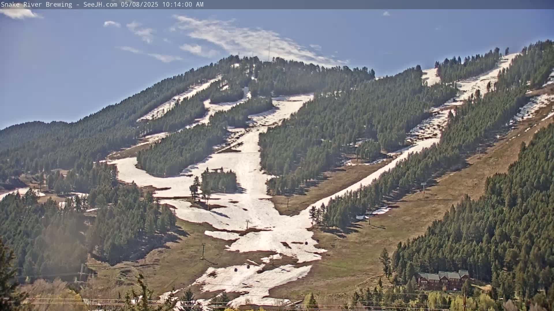 A mostly sunny day reveals a mountainside ski area with patches of remaining snow on the slopes and green grass in between, surrounded by evergreen trees.
