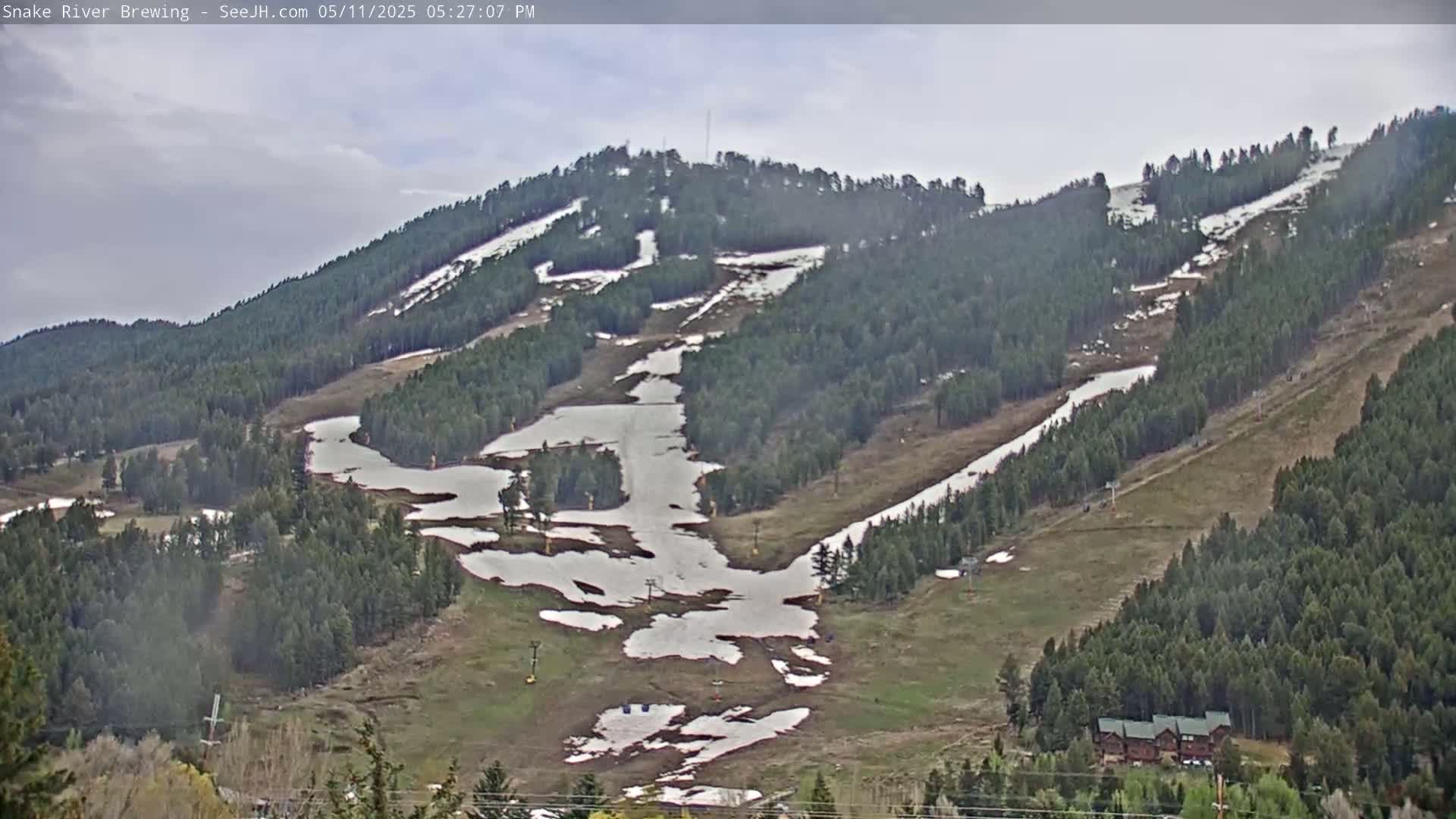 A mostly clear sky hangs over a snow-dusted mountainside with ski runs and a scattering of trees, patches of melting snow remaining on the slopes.