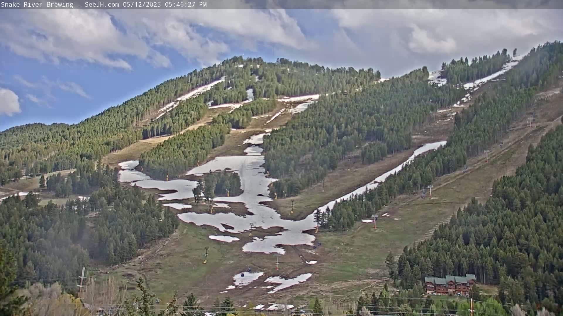 A partly cloudy day reveals a mountainside ski resort with patches of melting snow on the slopes and greening trees.